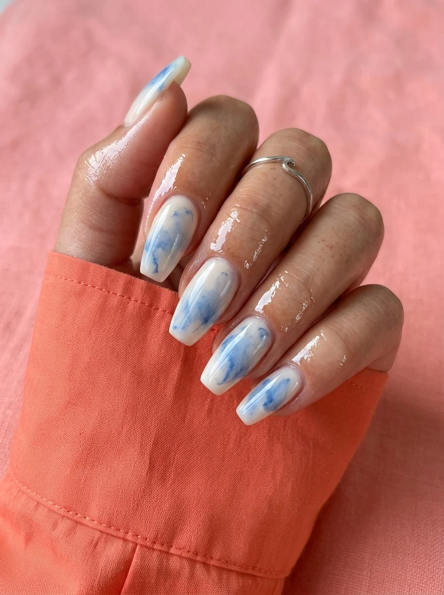 Almond nails with milky white base featuring soft blue watercolor marble swirls that look like ink dropped in water.