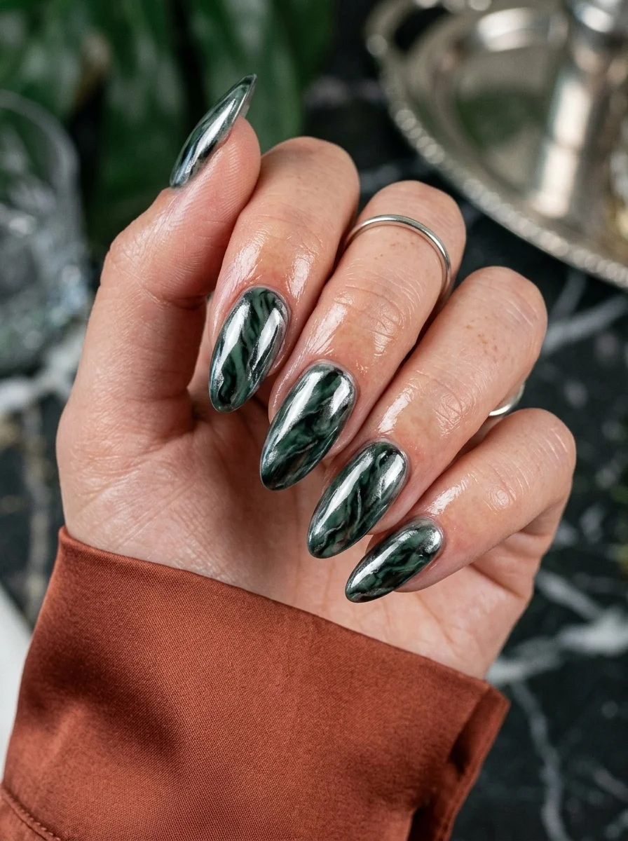 Almond nails with a dark green chrome base and silver or white marble veining throughout.