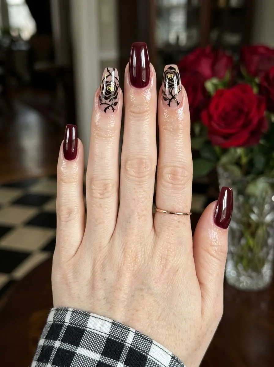 Deep burgundy coffin nails with sheer accent nails featuring black rose outlines and gold details.