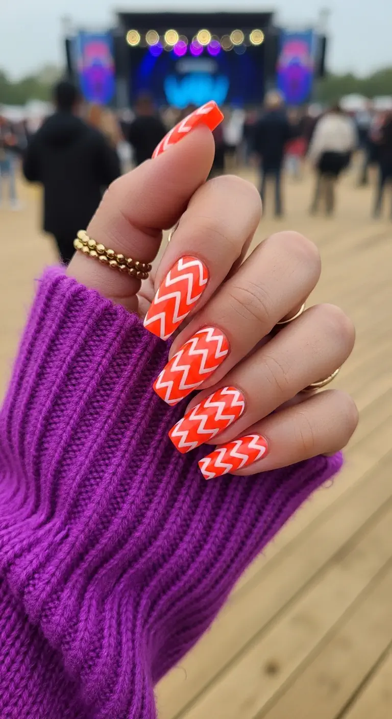 Long square neon orange nails with thick white zig-zag chevron patterns, shown at an outdoor music festival